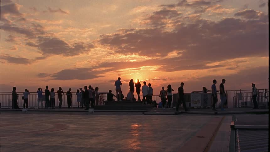 An audience of people standing against a railing looking at the sky where the sun is setting