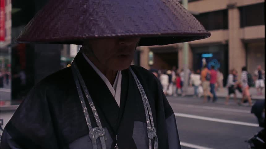 A japanese salesman standing in a busy street wearing traditional clothes and bambu hat as people go by