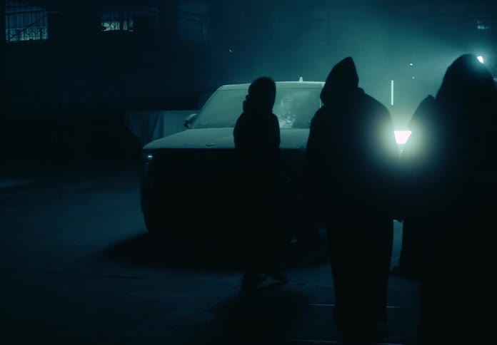 A group of hooded figures surround a pick up truck in a garage at night and one of them walks toward the truck while a light moves around it and its headlights turn on