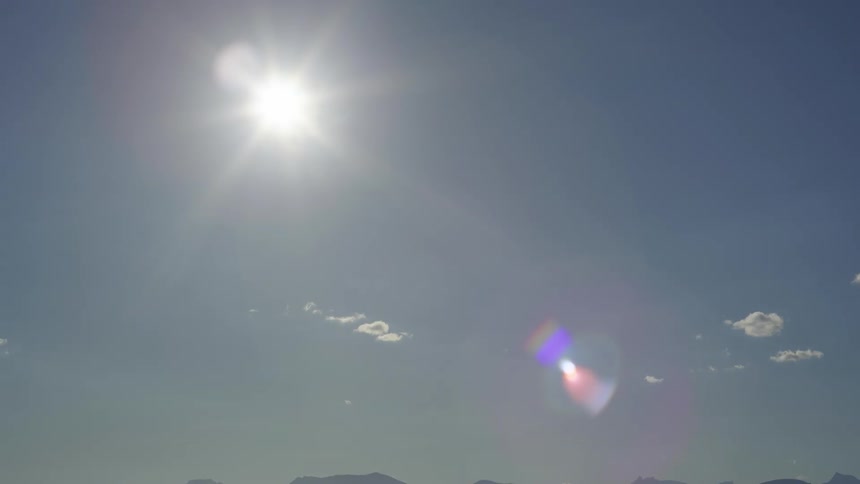 The sun shining brightly in the sky and there is a flare of lights and in the sky there are small white clouds meeting the blue sky