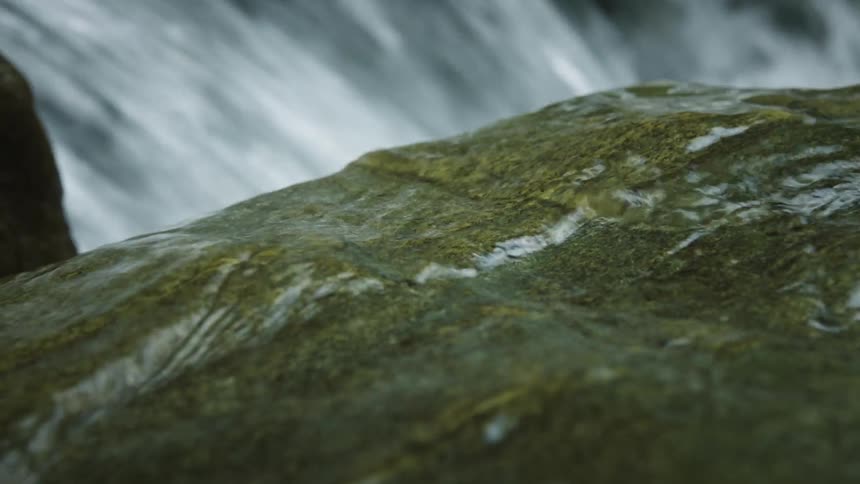 A closeup of a mossy rock with water flowing over it and a waterfall in the background