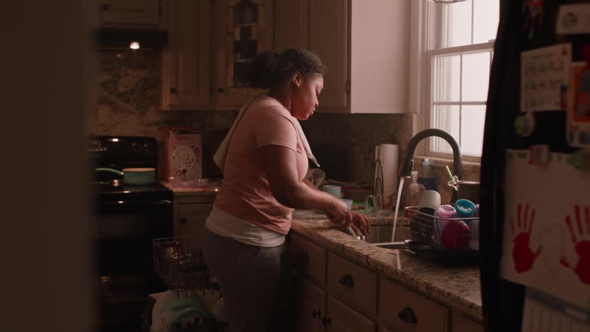 A woman is standing in front of a sink and is washing a woven dishware utensil while looking out the window