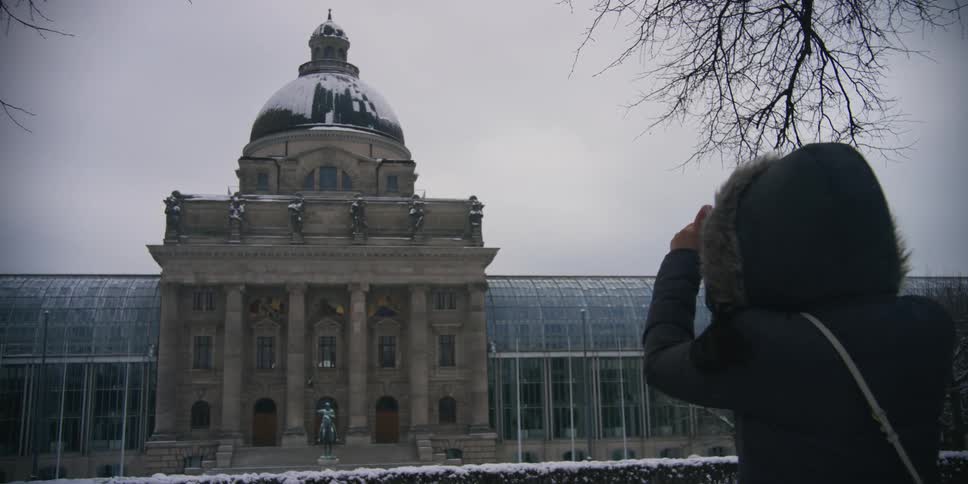 Back view of a woman in a jacket and hood pointing her cell phone at a grand historic building with a prominent dome and classical architecture on a cloudy winter day