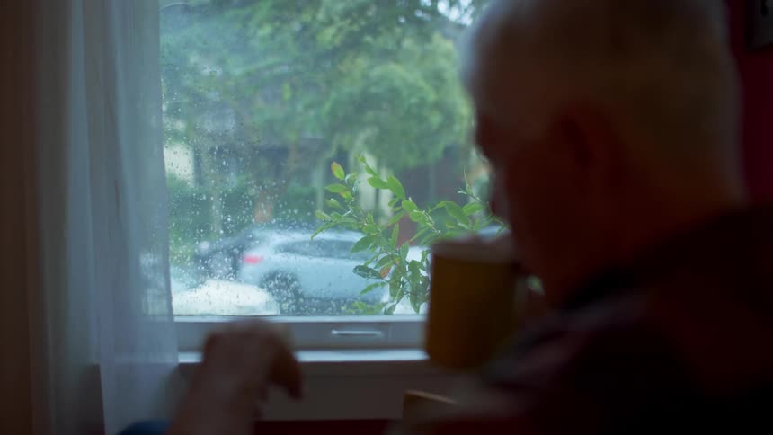 Elderly man sitting in an armchair drinking a cup facing the window with cars passing by on the street