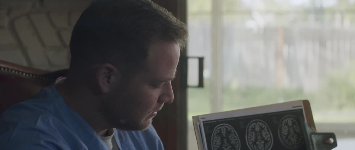 A doctor sitting in a chair holding a chart with a ct scan pointing at it with a pen while explaining it