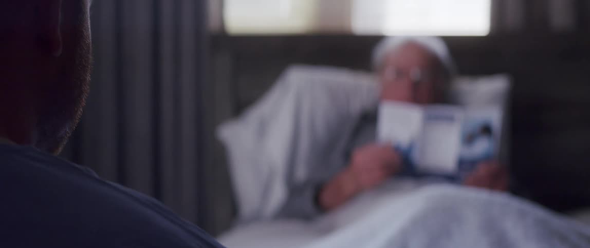 A stern elderly man resting in bed reading a brochure