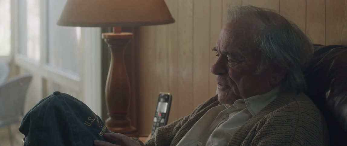 An old man in a chair takes off his hat talks about it then puts it back on