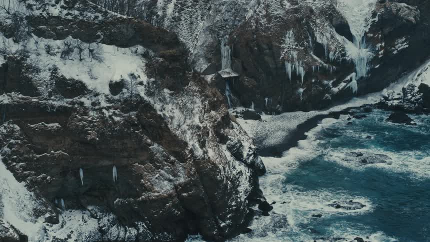 Aerial landscape of a crystal clear blue sea crashing onto the rocky shore covered in snow