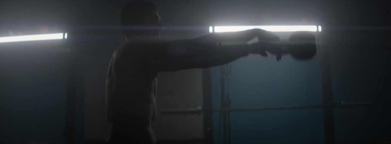 Man inside a gym with low lighting squatting and lifting weights that throw from one hand to the other