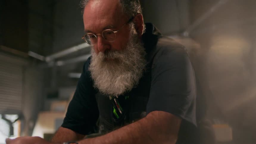 Bearded man sanding a slab of wood in a workshop