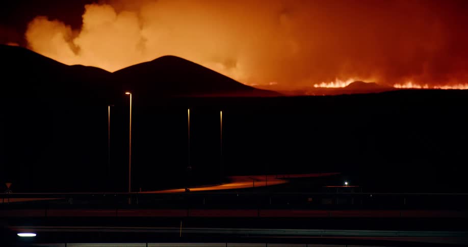 Glowing red smoke fills the sky as flames rise in the distance and traffic passes on the road