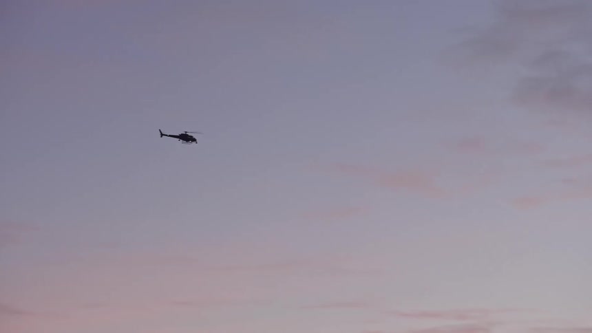 A helicopter flies across the open sky as tail lights flash