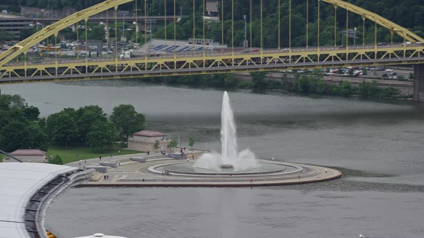 A large outdoors fountain is located on the waters edge near a bridge and sports stadium