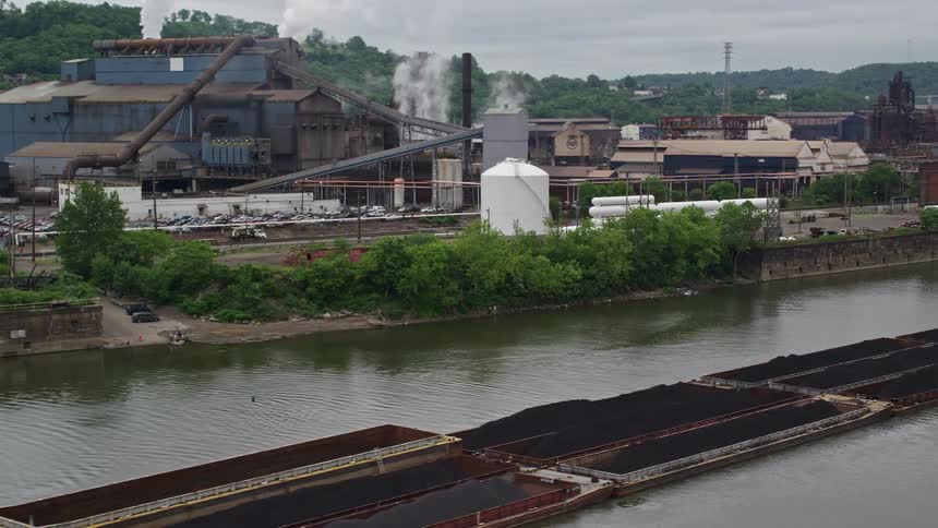 Tug boat pushing coal on a river beside a steel factory with smoke bellowing from its stacks