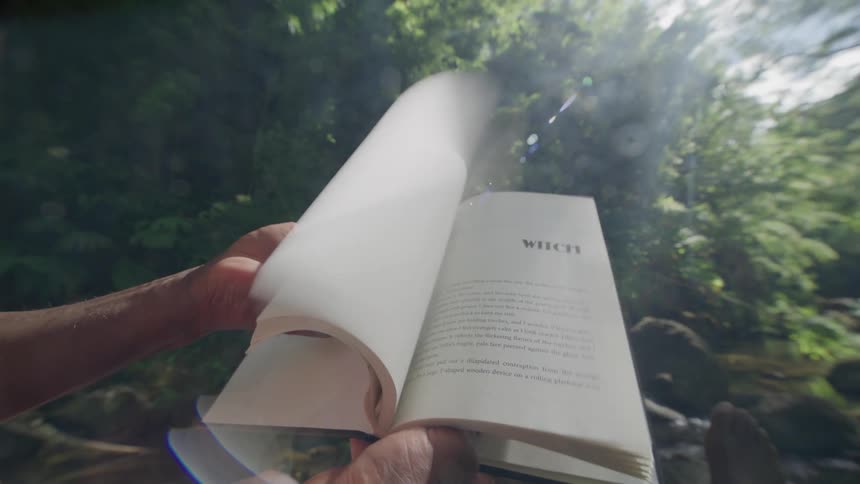 Reader flips backwards through the pages of their book in front of trees and a stream