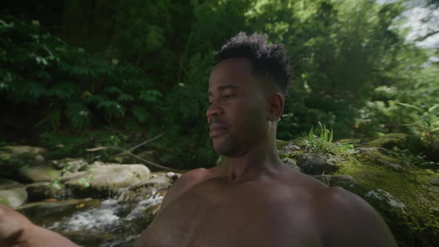 Man without a shirt sitting on a rock next to a river reading a book