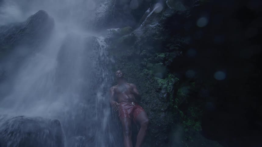 A fit man stands under a waterfall surrounded by rocks