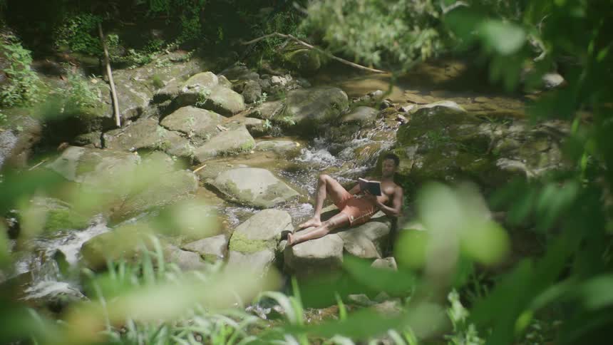 A man relaxes on some rocks beside a creek as he reads a book