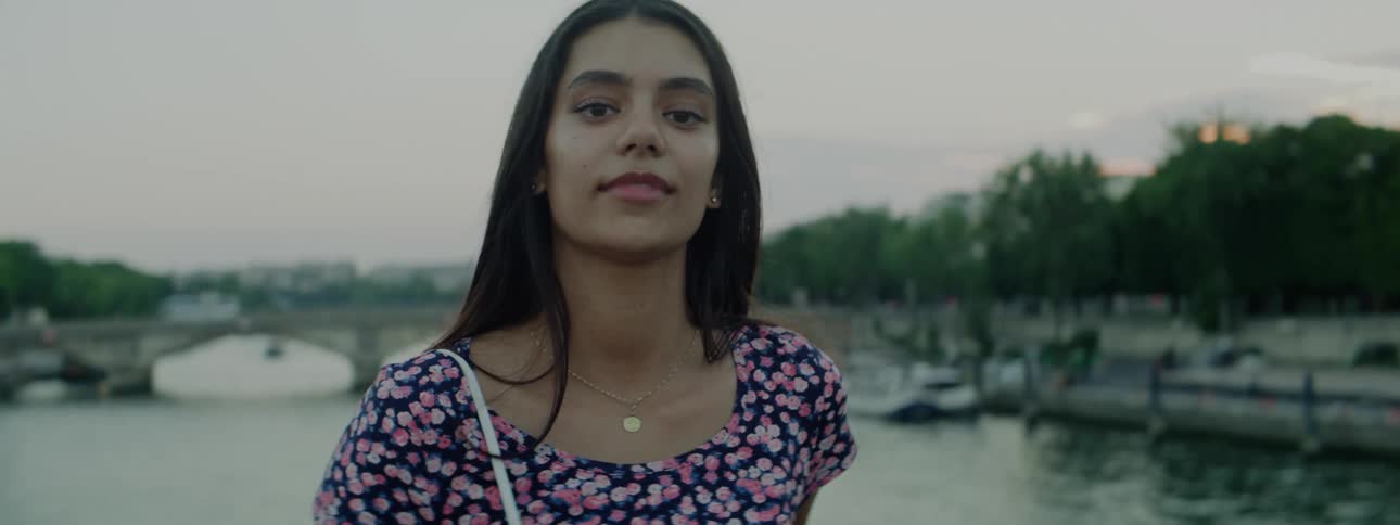Woman brushes her hair from her faces as she talks in front of a bridge crossing a river
