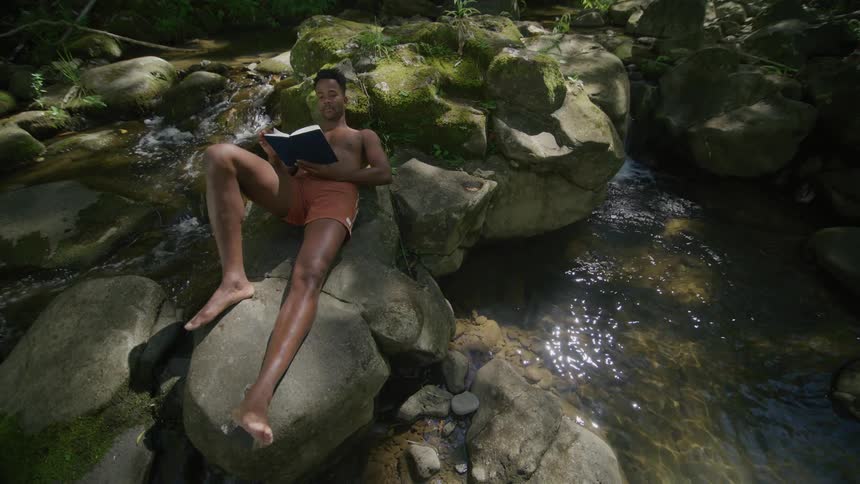 Man in shorts lying on a rock reading a book in a river