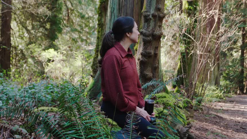 A young woman sits in a forest drinking out of a travel mug