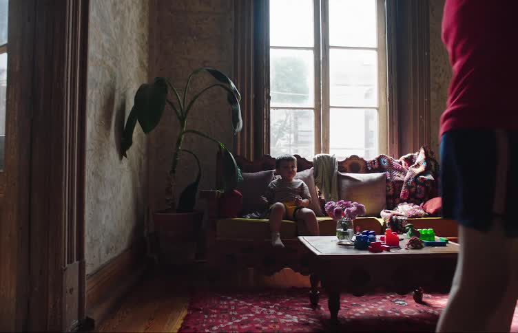 A boy sits on the couch while his sibling dribbles a ball indoors