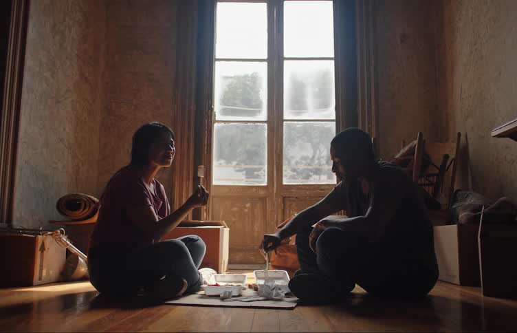 A man and a woman are sitting on the floor next to a window while they are eating and talking