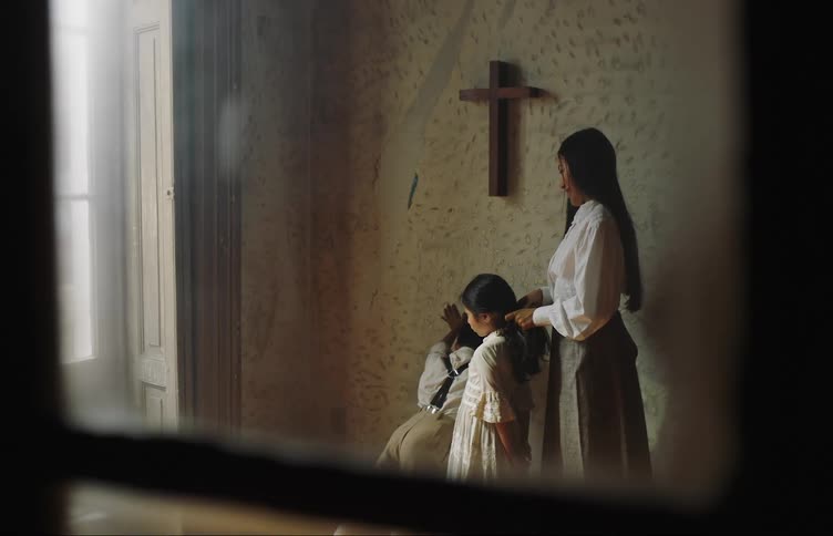 A woman braids a young girls hair next to a wall with a crucifix while a young boy waits and moves around impatiently