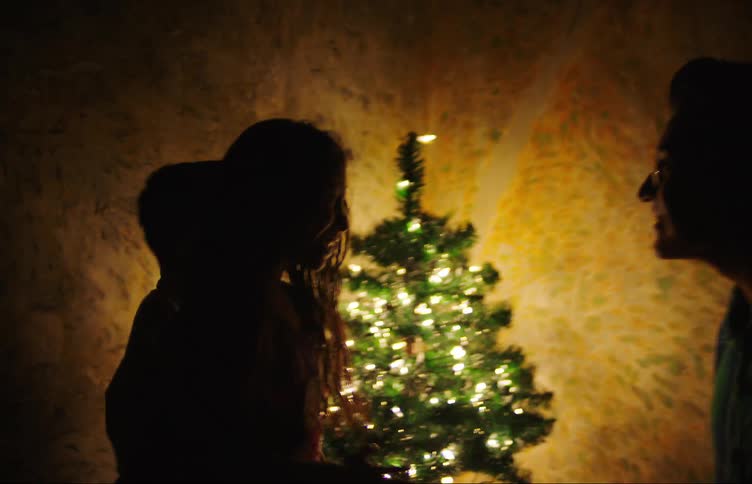 In a dark room a man calls two children to the corner where a christmas tree shines full of lights and gives them a gift