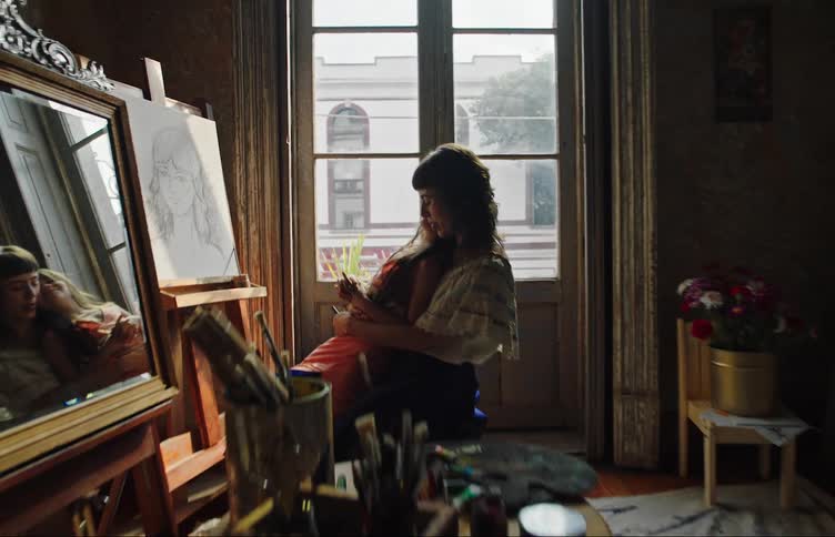 Mother sitting with her daughter on her lap while they are facing an artistic white canvas painting in a room with paint frames