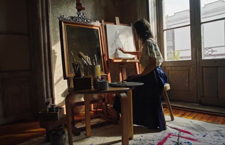 A woman sitting at an indoor easel sketches a profile on a canvas