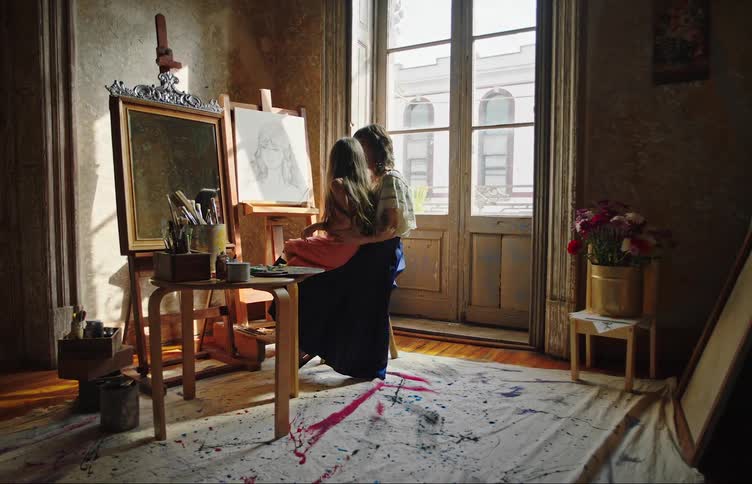 A girl sits on the lap of a woman painting in a studio
