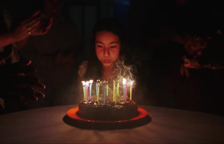 Portrait of a young birthday woman sitting at a table while her colleagues around her sing happy birthday and she then blows out the candles and everything goes dark