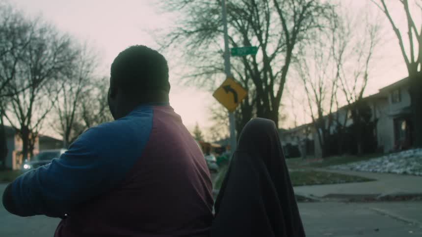 A man in a wheelchair is waving at a yellow bus that passes straight down the street