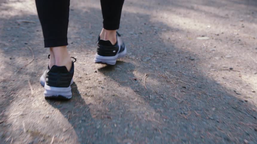 The legs of a person wearing black pants and sneakers is walking forward on a gravel trail