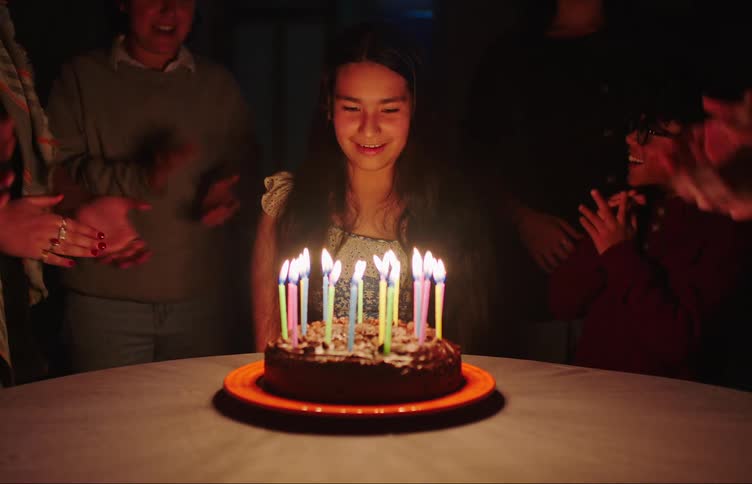 Teenager sitting at a table in front of a cake with birthday candles and her family around her clapping and then blowing out the candles