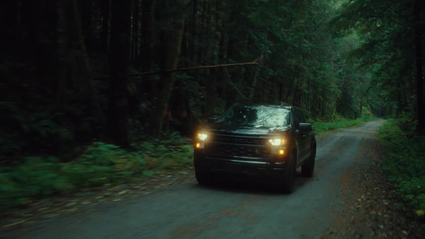 Black pickup truck with headlights on traveling along a narrow road in the middle of the forest
