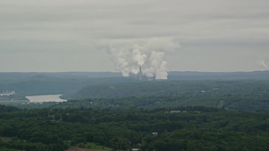 Puffy clouds of smoke rise from smokestacks in the distance behind a vast forest