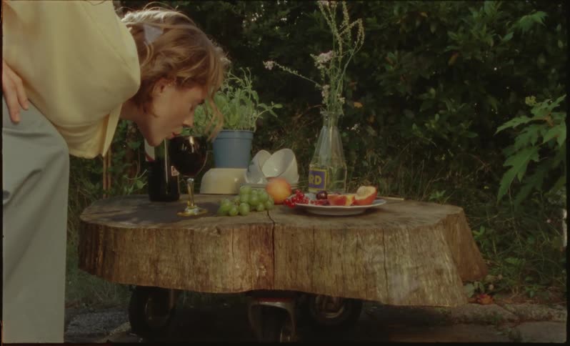 Young woman in a garden crouching to take a sip from a glass of wine next to fruit