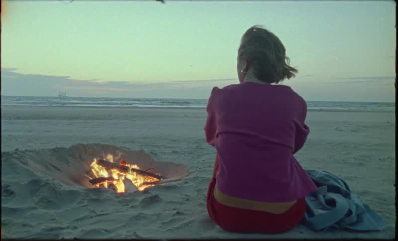 Back view of a woman sitting on the beach next to a bonfire on a late afternoon with a sea breeze