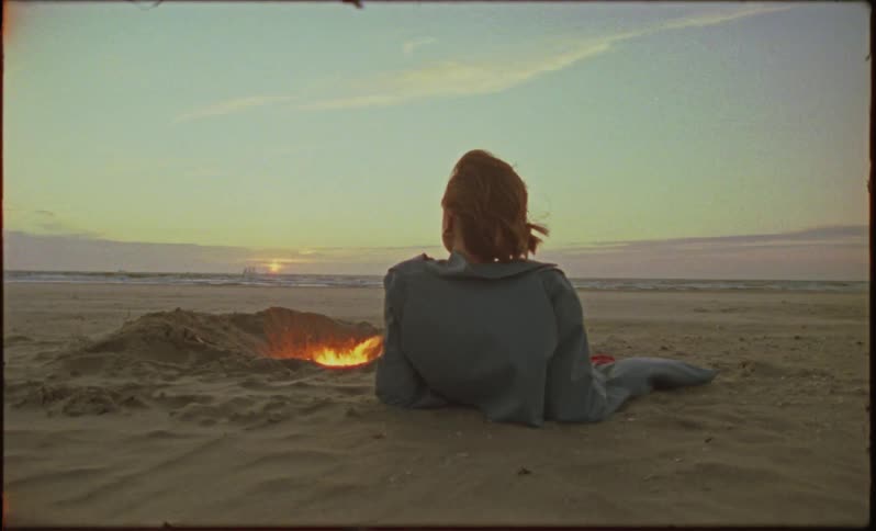 A woman lays on a sandy beach with a fire burning in a pit next to her
