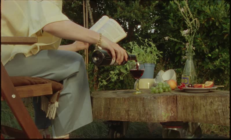 Woman sitting on a bench facing a wooden countertop and pouring wine from a crystal glass