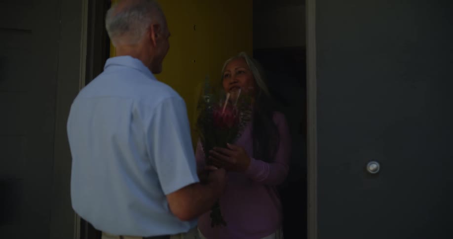 Elderly man being greeted by a woman who opens the door of the house and hands him a bouquet of flowers they greet each other and enter
