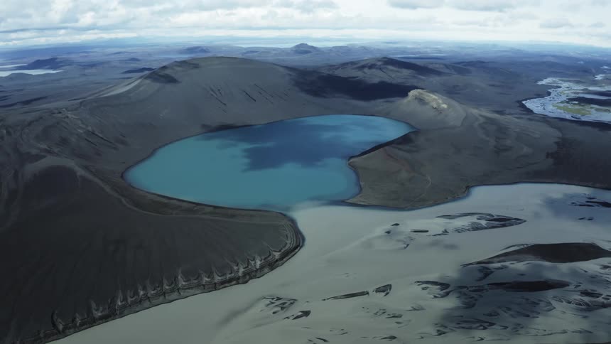 An aerial view of a turquoise lake nestled between dark mountains under a cloudy sky
