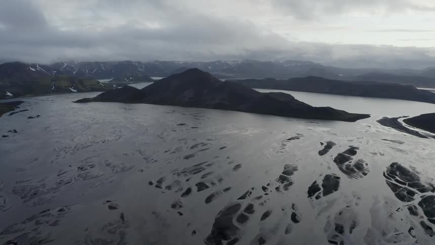 Aerial view of glacial crater lake with braided river and large central rock formation under low clouds