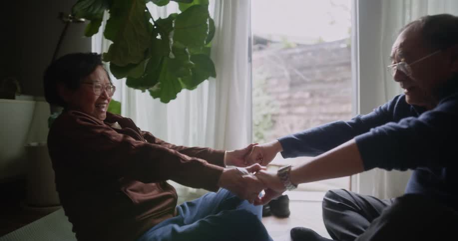 Elderly couple sitting in the living room and on the floor holding hands stretching each others bodies