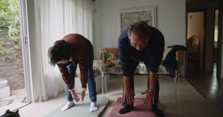 An elderly couple does yoga stretches and breathing exercises in their living room