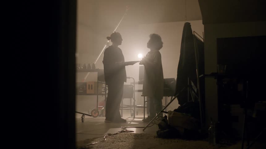 Two people standing side lit by a glaring light have a conversation