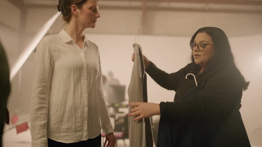 Two women helping another woman wear a coat while talking in a studio