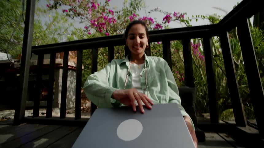 A young woman sits outdoors on a wooden deck while opening up her laptop